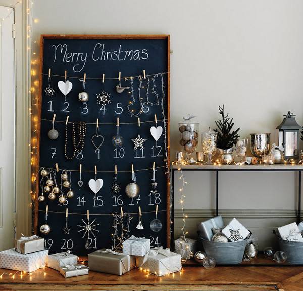 Chalkboard converted into advent calendar with hanging ornaments on string with clothespins near table decorated with Christmas decorations.