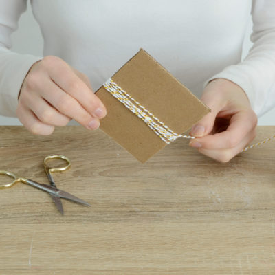 Person is holding a cardboard tied with a thread and scissor, thread bundle on the table.