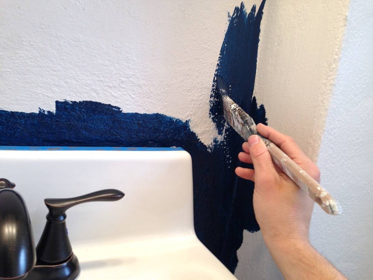 A hand holds a paintbrush, painting the white wall above a white sink blue.