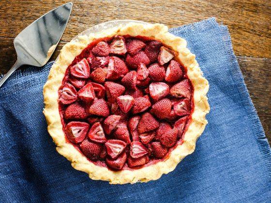 Pie shell filled with sliced strawberries and a serving utensil, sitting on a blue cloth.