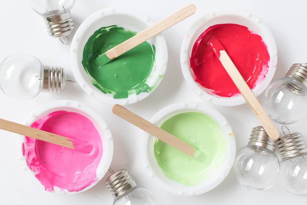 Four bowls with different color paste of red, dark green, lime green and hot pink with popsicle sticks in them and light bulbs surrounding them.