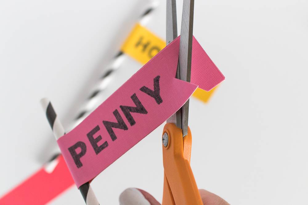 Orange handled scissors cutting through a small piece of pink paper with black lettering on it.