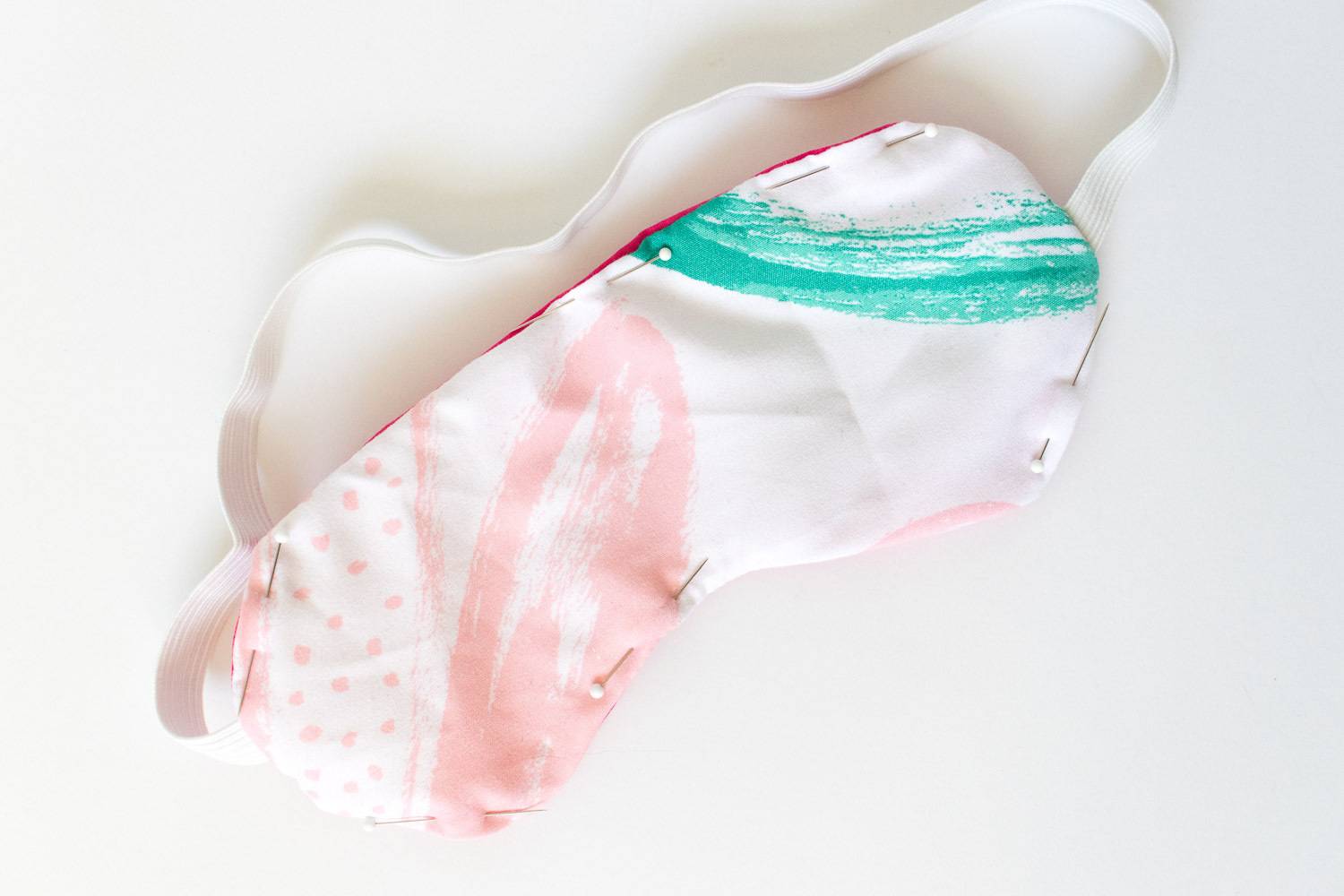 Cotton eye mask that has colors white, pink, and green with a elastic strap.