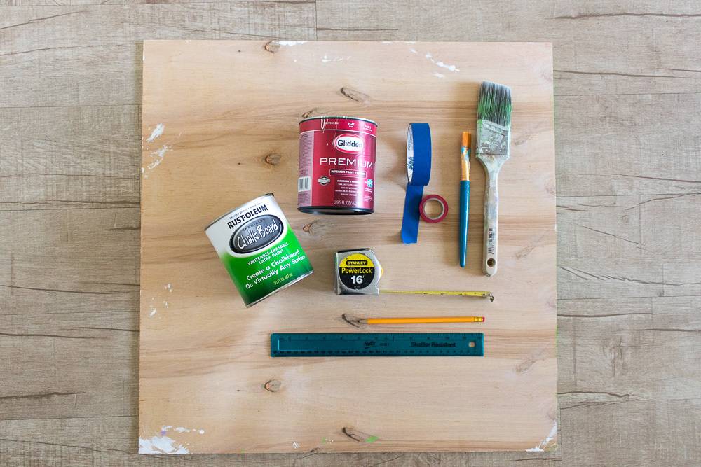 Paintbrushes, paints, tape measure, ruler, pencil and other tools atop an unfinished square wooden board.