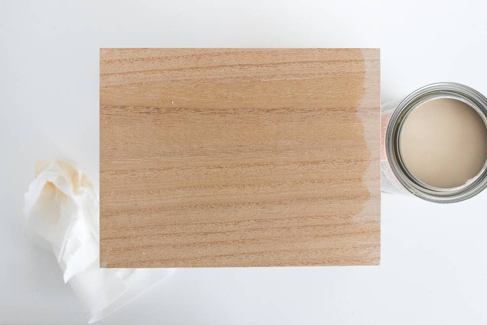 Open can of neutral-colored paint next to a squarish unfinished wooden board.