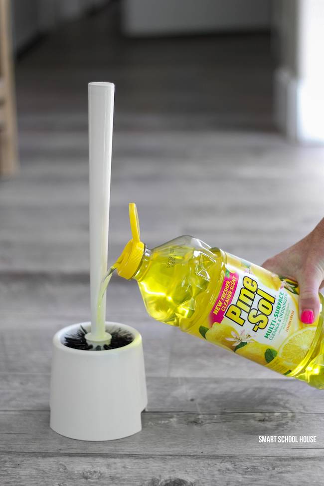 A person is holding a bottle of yellow cleaner near a toilet brush.