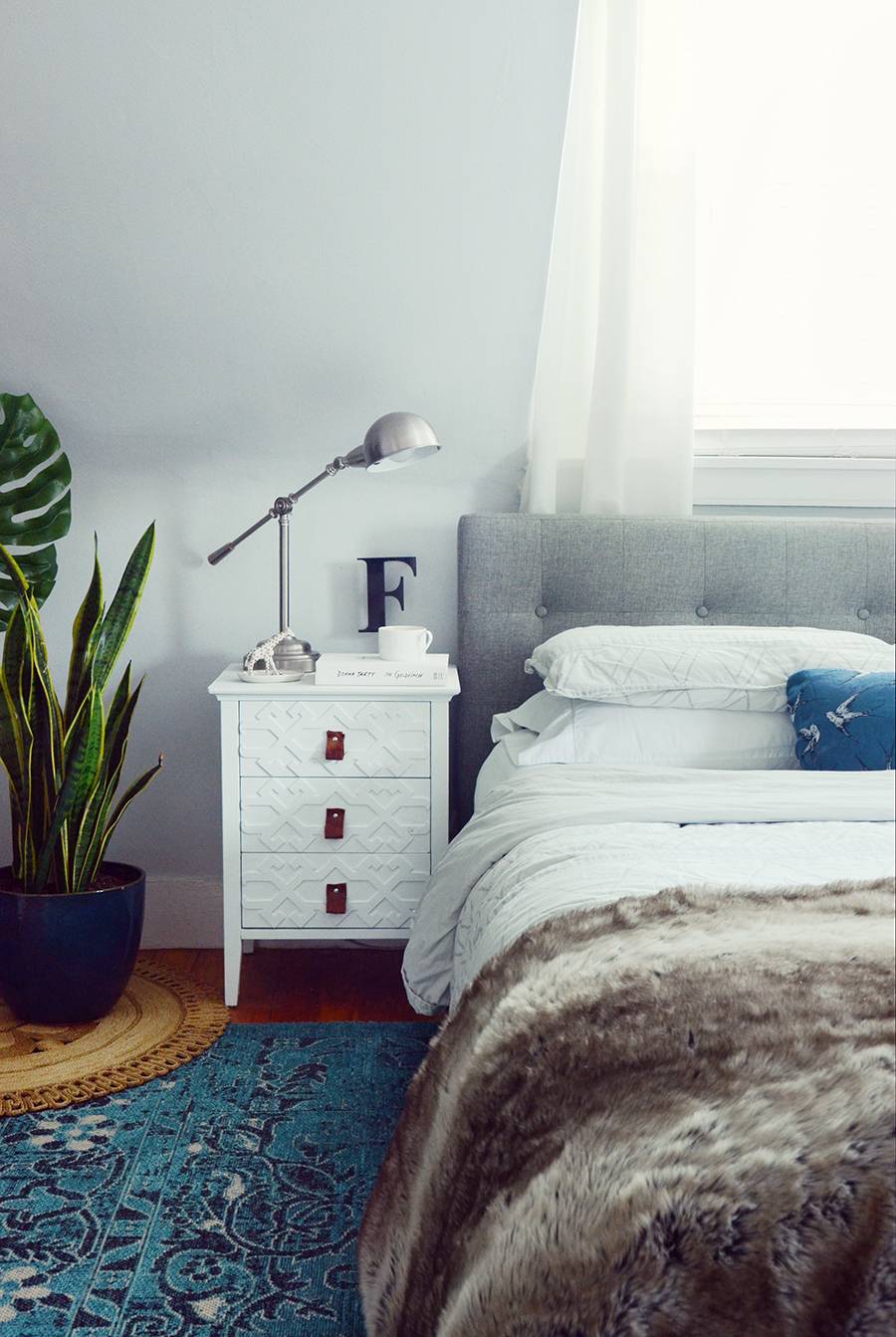 A lamp is sitting on a white bedside table between a bed and a plant.