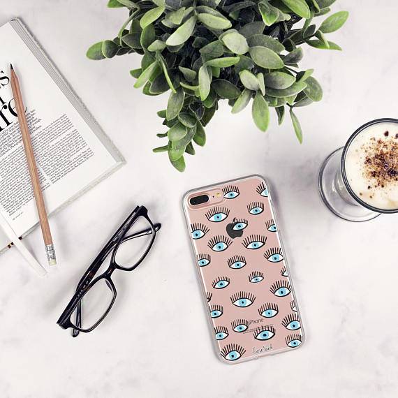 An open book with a pencil on it laying next to a pair of black readers which are laying next to a cell phone with a pink case full of eyes with long eyelashes on it which is next to a glass cup of frothy coffee.