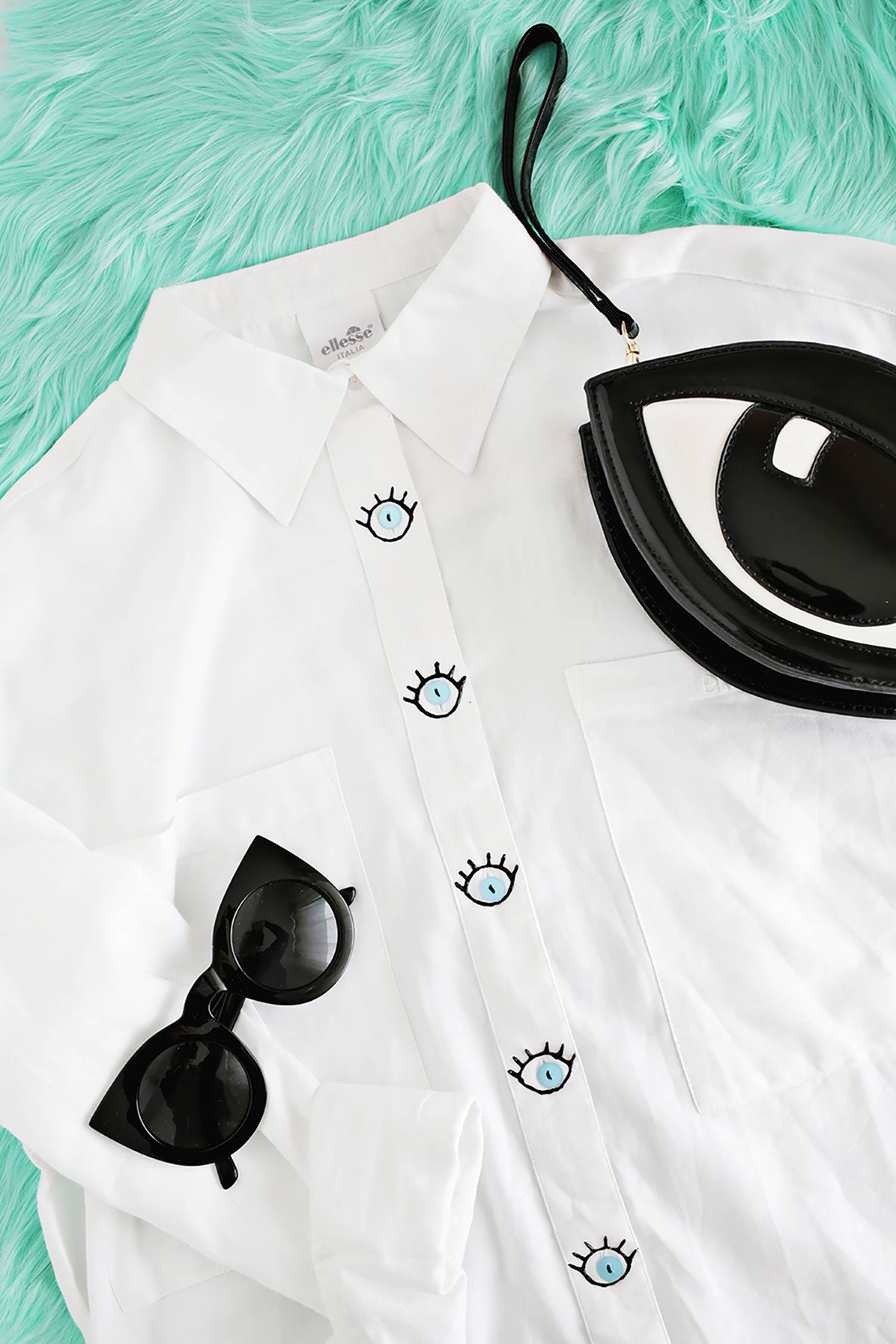 A white button up shirt with eye ball and eye lashes for the buttons, a pair of black sunglasses and a black handbag shaped like an eye.