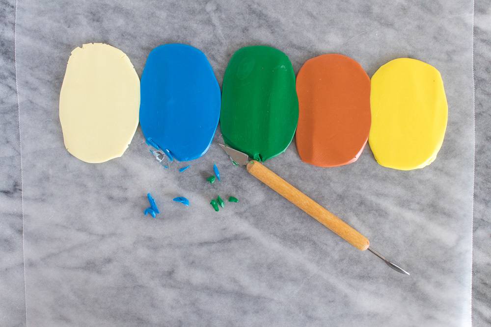 Five vertical ovals of colored clay, one white, one blue, one green, one orange and one yellow above a wooden craft cutter.