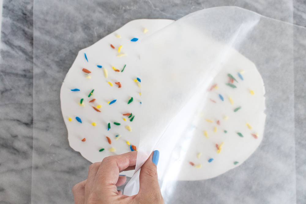 A woman's hand with blue nailpolish is pulling a piece of wax paper over a white rolled out piece of dough with multi colored pieces spread over it.