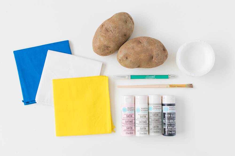 Blue, white and yellow napkins next to two brown potatoes, a white cap, two paint brushes and four small containers of paint.