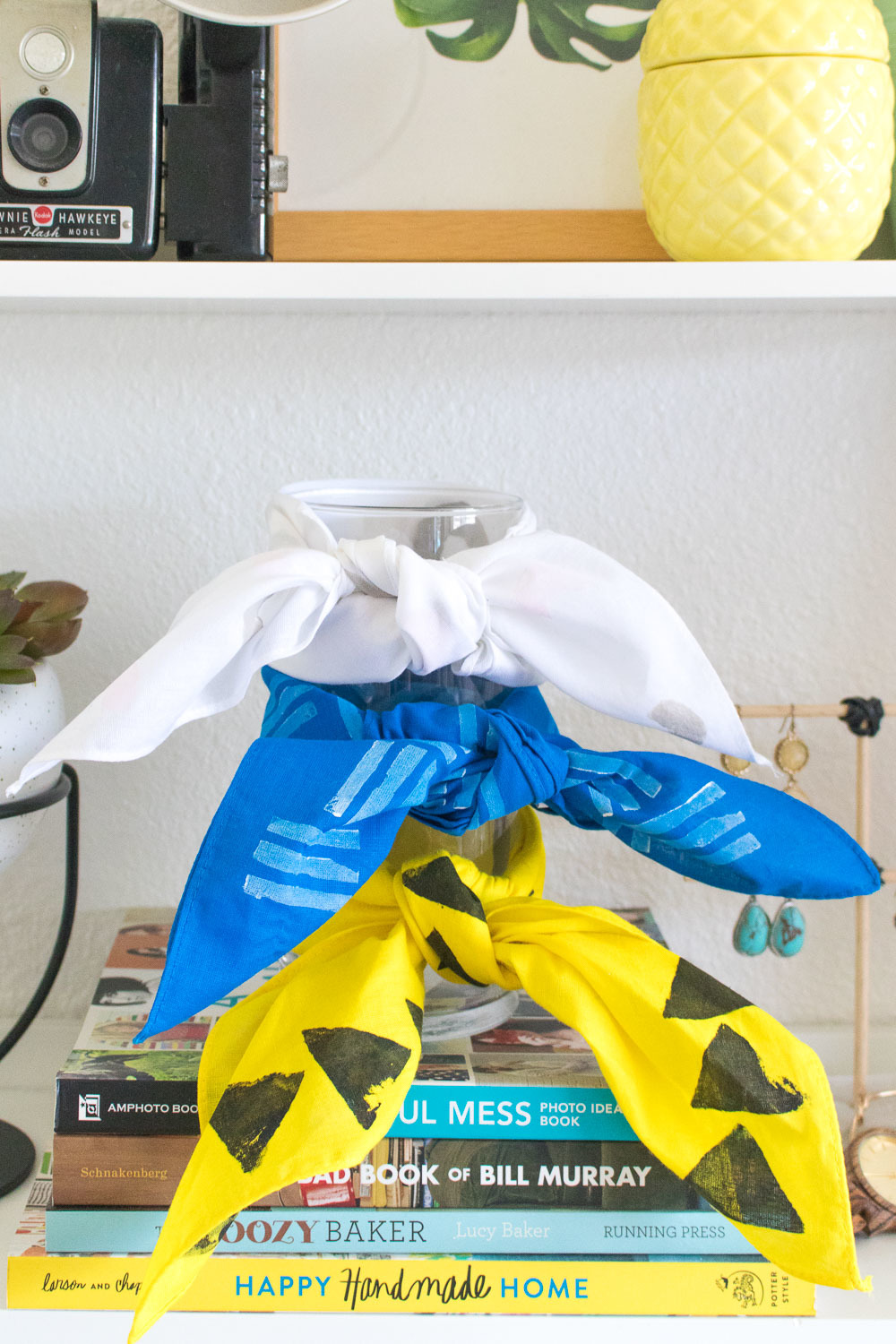 Three pieces of cloth tied around a glass vase, one white, one blue and one yellow and black all sitting on top of a pile of books.
