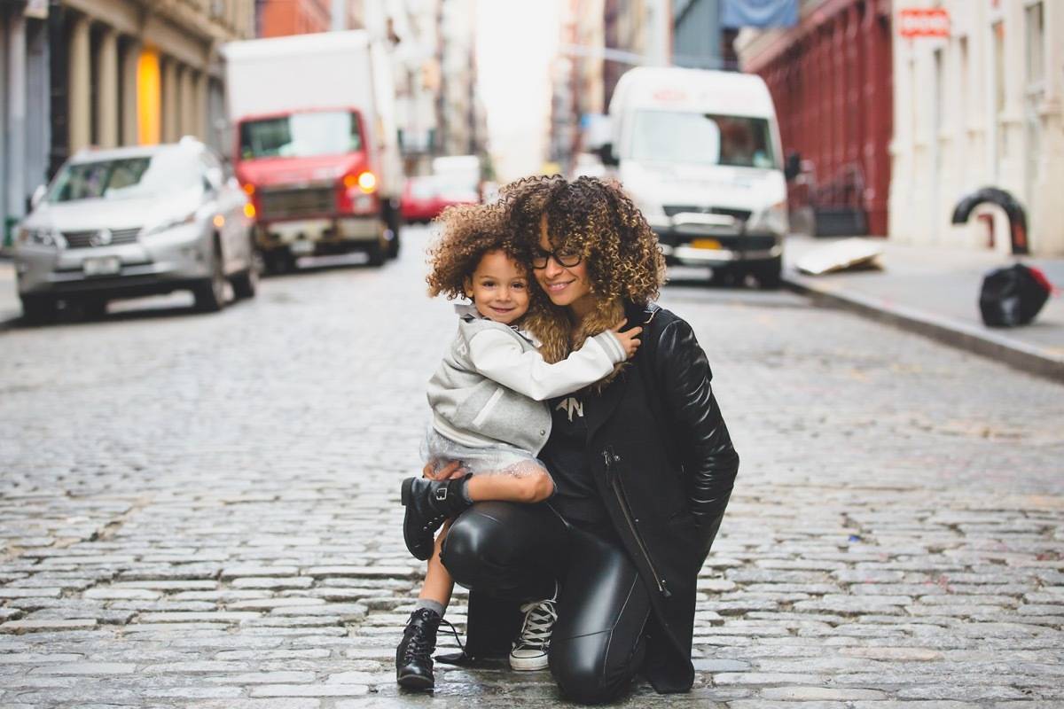 A black lady a black leather kneels in the street hugging her child in a large city.