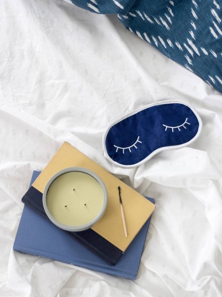 A candle on top of two books next to a blue sleep mask on a white sheet.