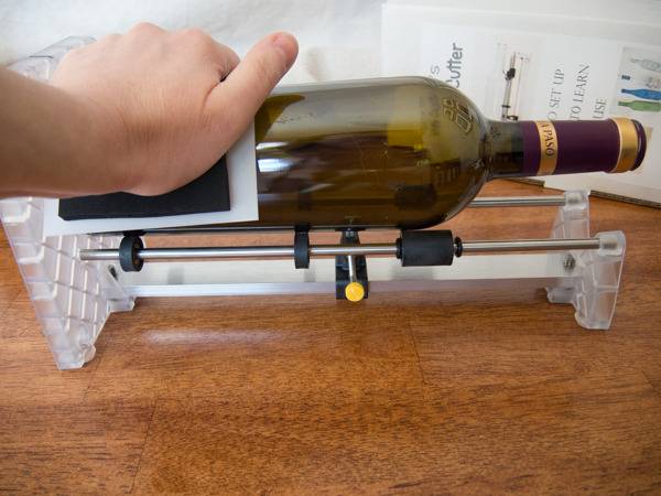 Hand holding a horizontal empty glass wine bottle on a bottle cutter, on a wood surface.