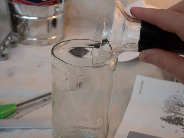 A person holds up the neck of a glass bottle over the bottom part of the glass which has fluid in it.