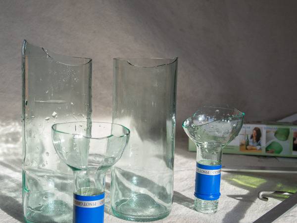 Two glass bottles are broken about a quarter of the way from the top, with the tops sitting upside down in front of the bottom of the bottles.