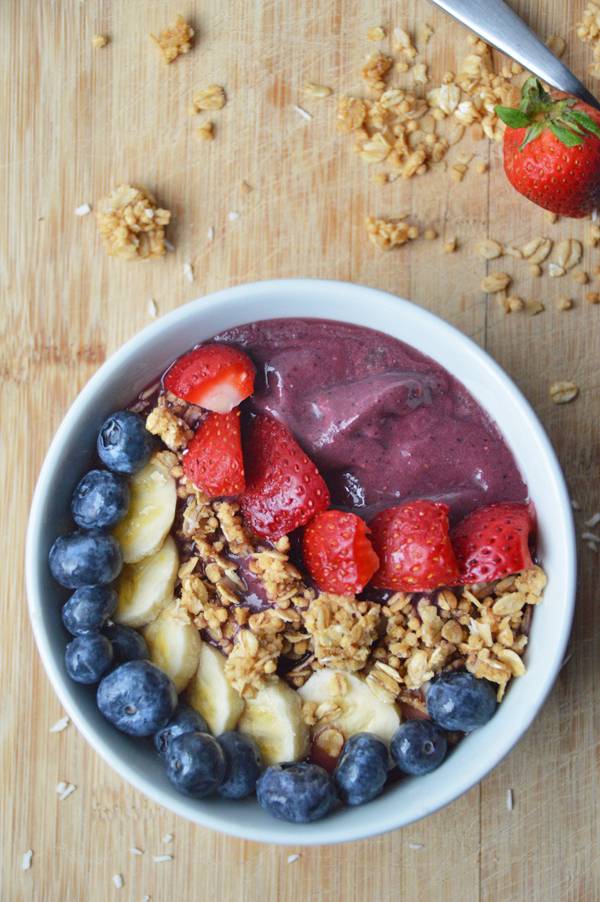 A white bowl of purple yogurt, red strawberries, brown granola, white bananas and blue berries all lined up.