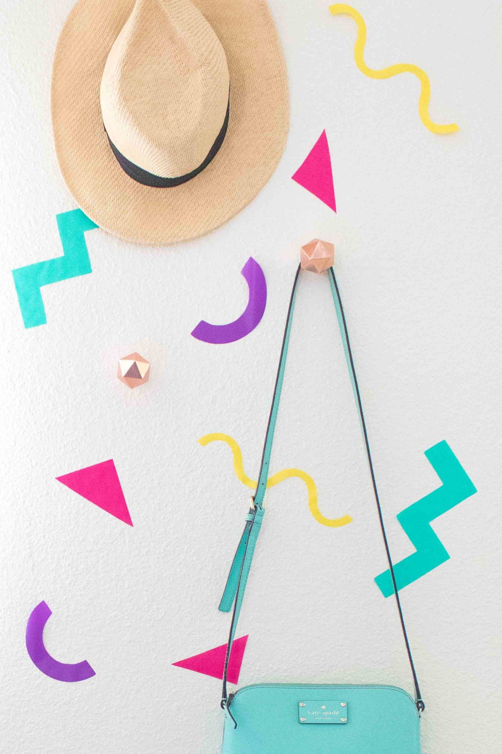 A purse and hat hanging on a white wall with some colorful decorative designs.