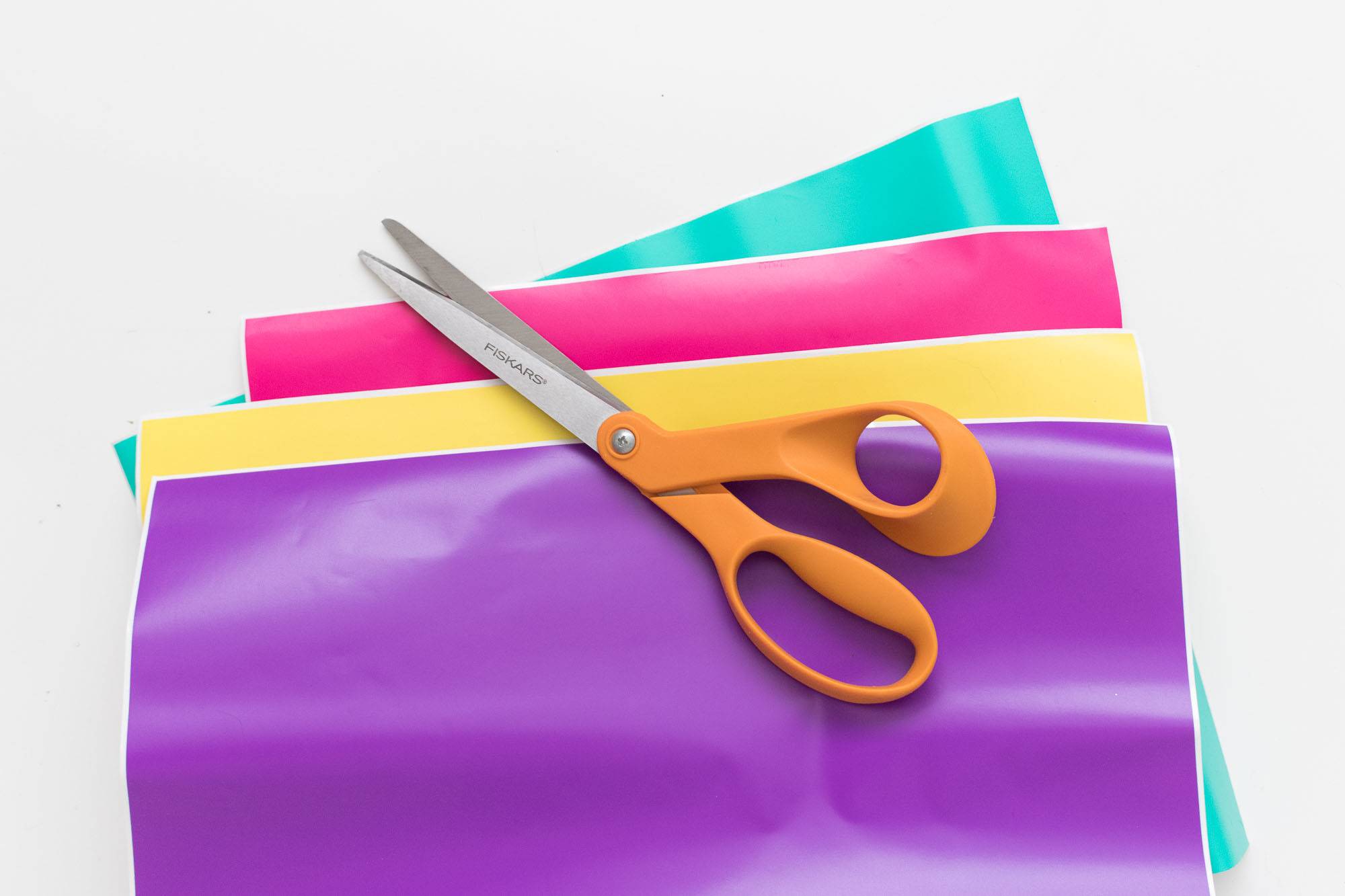 Orange handled scissors sit on different colored sheets of material.