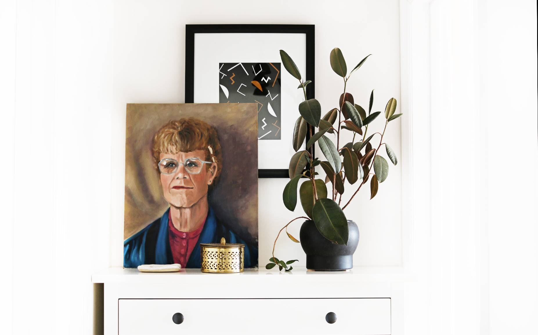 On a white chest of drawers, there is a potted plant on the right and a painting of a person to the left, with another picture visible behind it