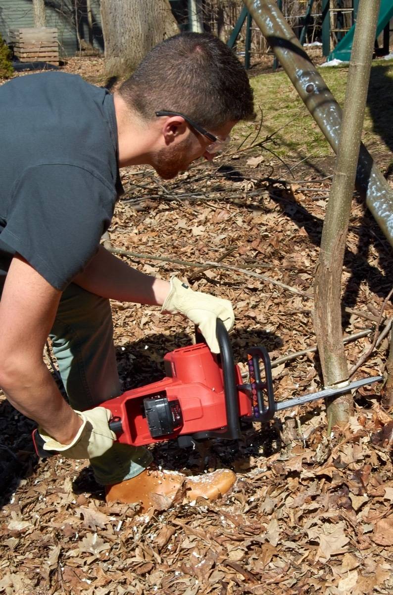 Man in safety glasses and gloves bending down to cut a small tree trunk using a chain saw.