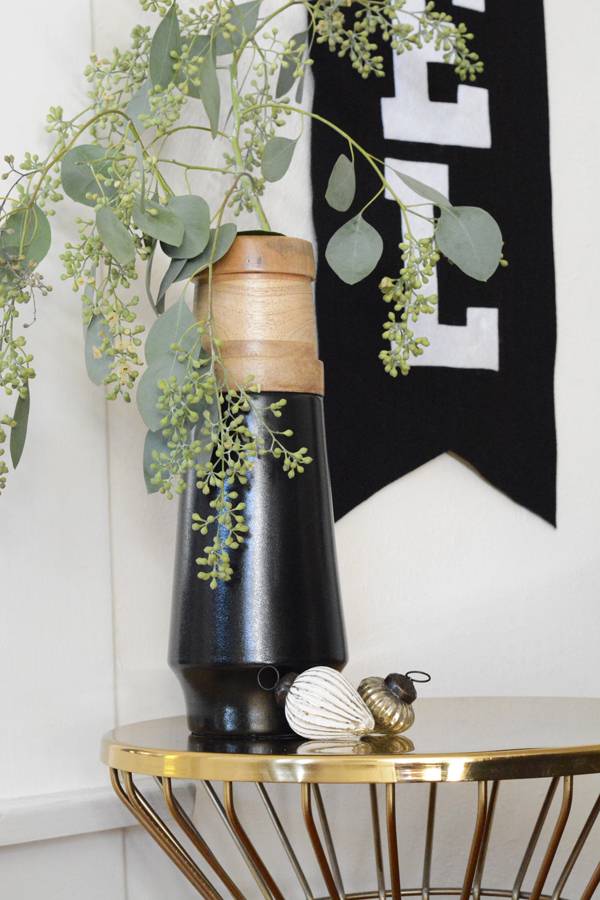 A round gold wire side table with a tall black vase with some eucalyptus leaves in it and a black banner hanging on the wall.
