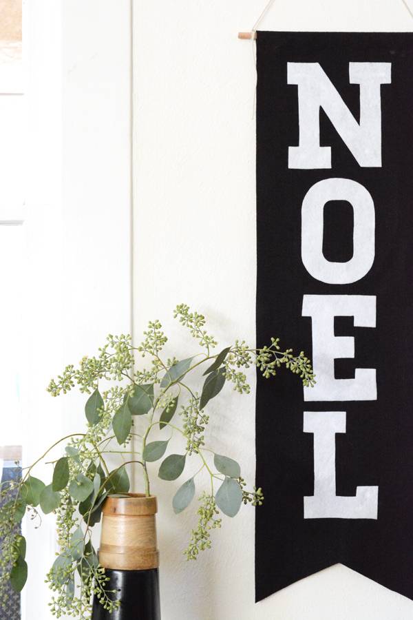 a Noel sign hanging on a white wall, next to a green flowering plant and a bottle of wine.