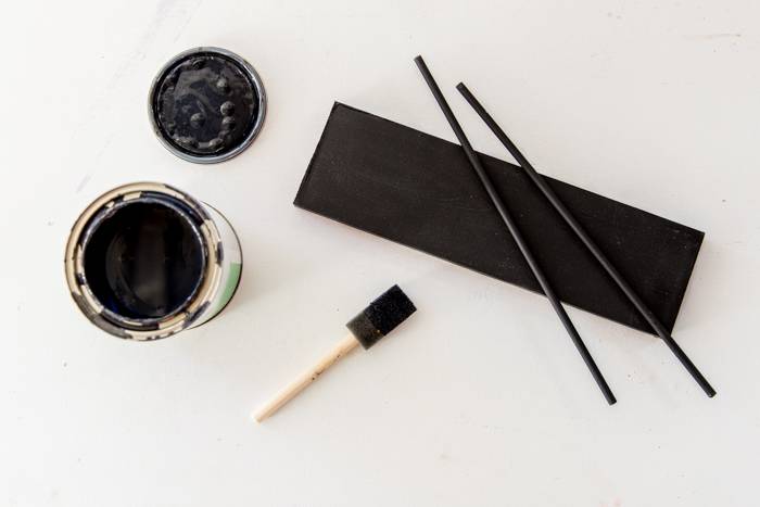 Black paint in a open tin container, paint brush and a black painted wooden block with two sticks.