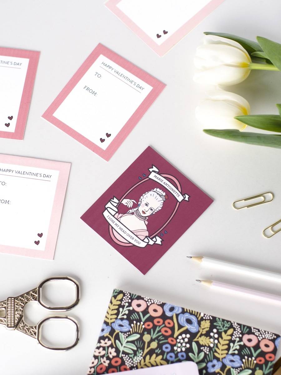 Valentines day cards sitting on a white table next to white roses and a notebook with flowers on its cover.