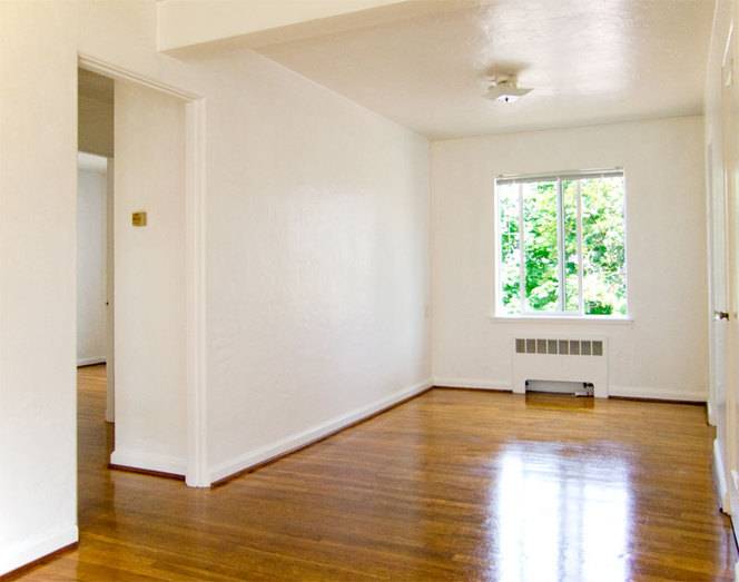 A bright empty room with white walls and glossy wooden floors, and a single window.