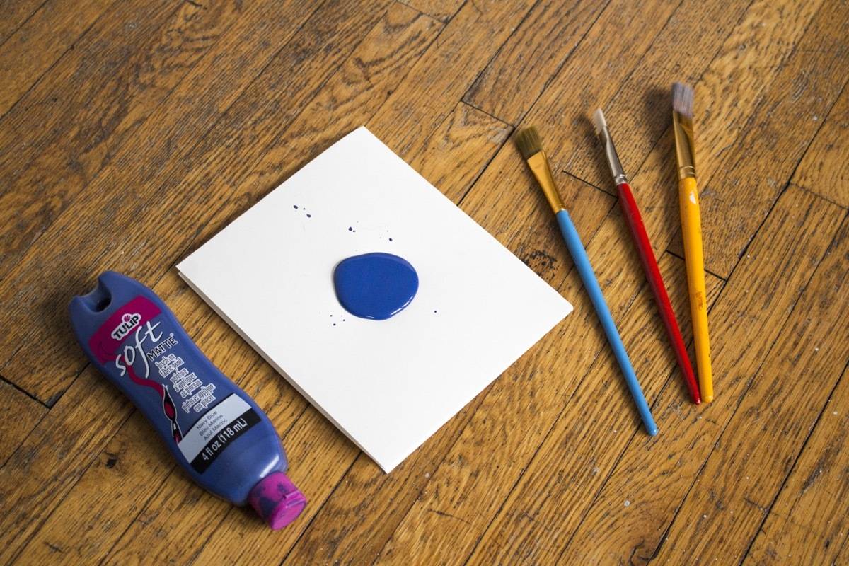 A white paper with blue paint is sitting next to the tube of paint and three paint brushes.