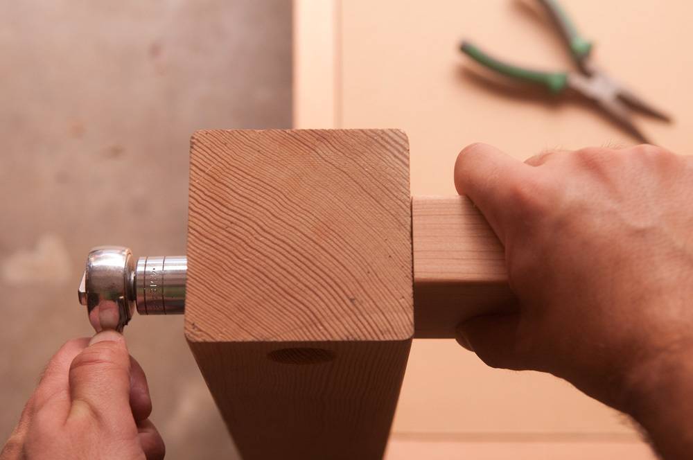 One hand using a tightening tool while the other grasps a piece of wood held next to a square upright beam.