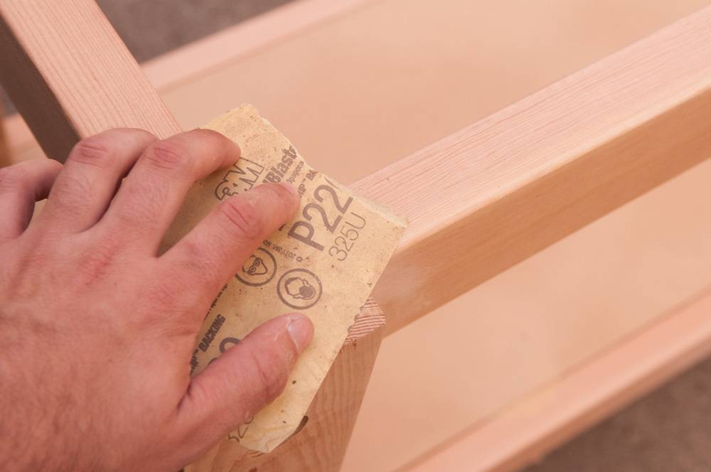 Hand applying sandpaper to polish the corner of a raw wood structure.
