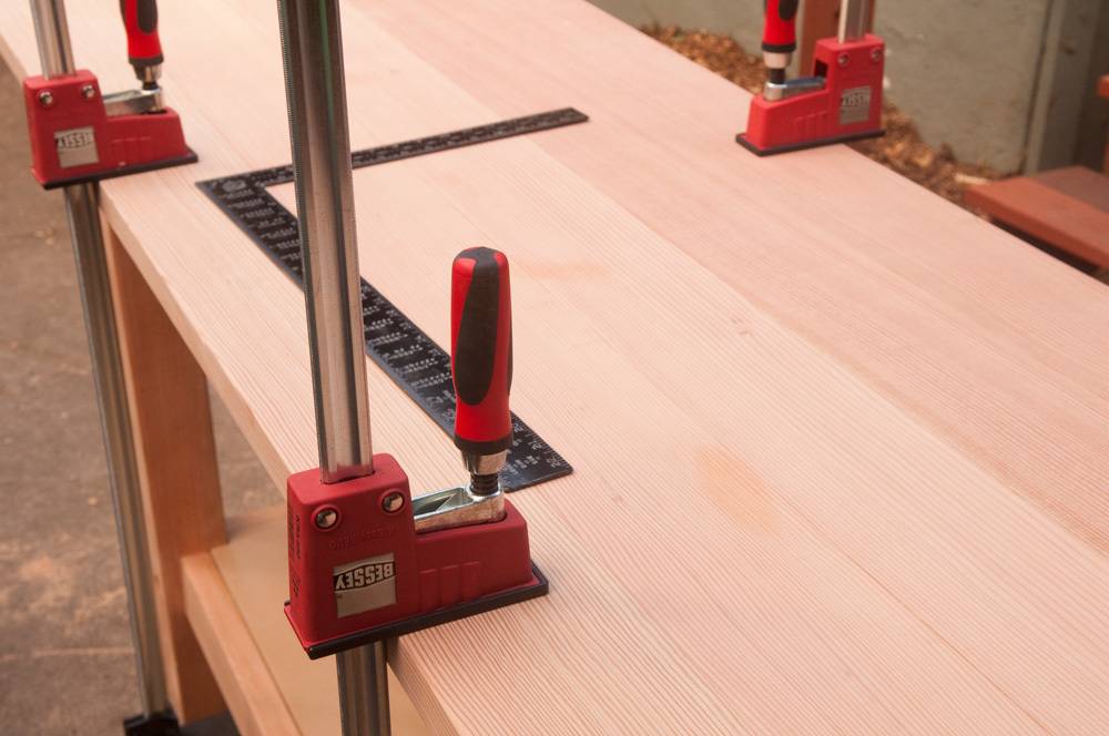 A red clamping device is holding a silver pole to a wooden surface.