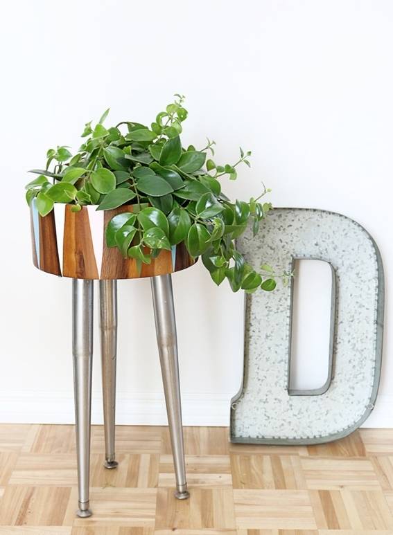 A sleek, three legged plant stand filled with ivy stands on a wooden floor beside an oversided metal letter D.