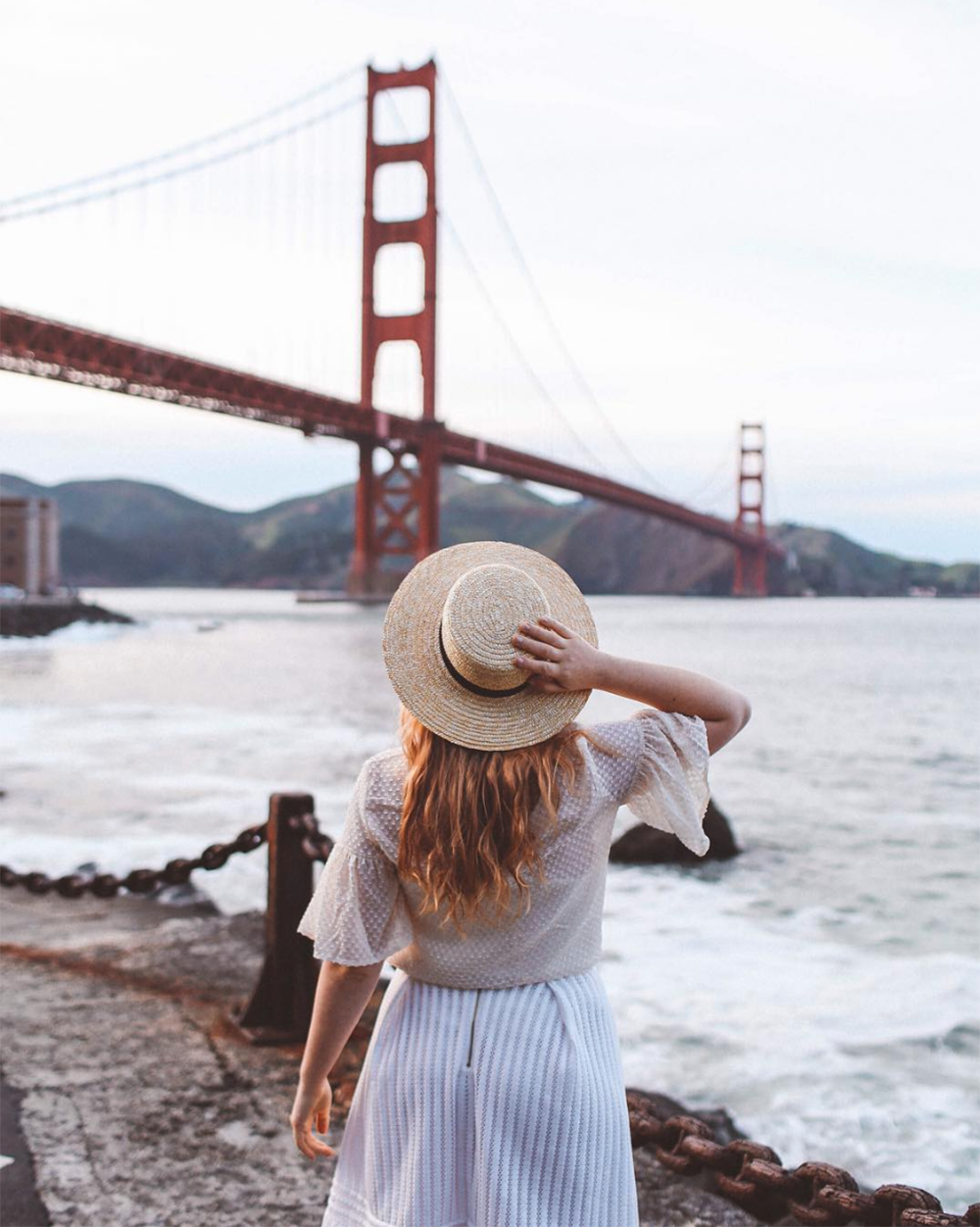 A woman in a hat is looking out at the water near a bridge.