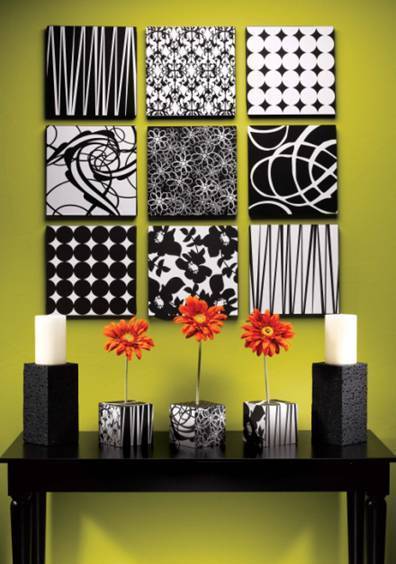 Nine square black and white pieces of art each with a different pattern are on a green wall behind a black table that holds two pillar candles and three short vases each holding a red flower.