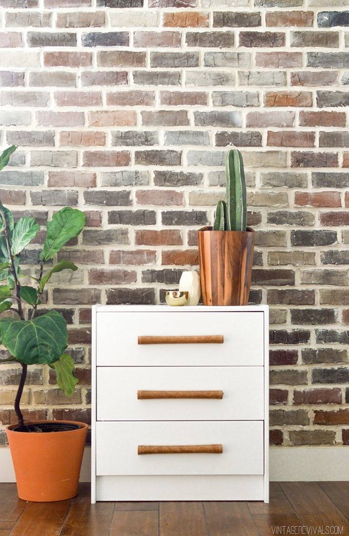 A white sideboard has a vase and is sitting in front of a brick wall.