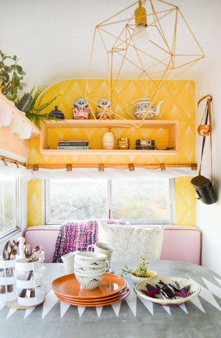 Inside a camper with yellow shelves a pink couch and a grey table with orange plates on it.