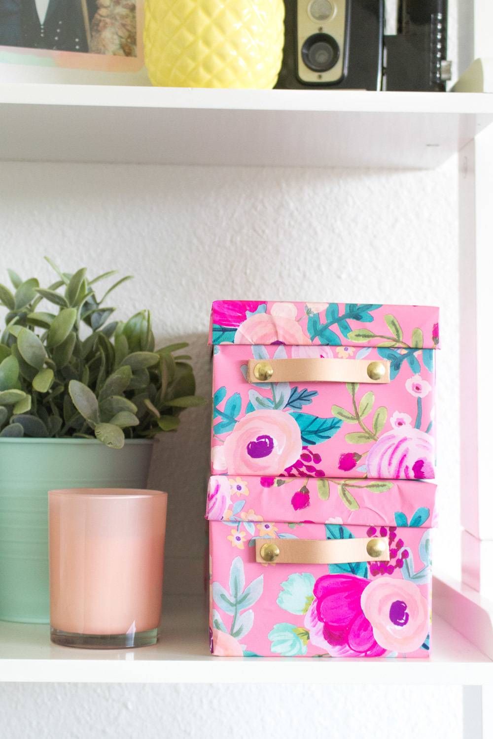 A white shelf has pink colorful boxes, a candle and a plant.