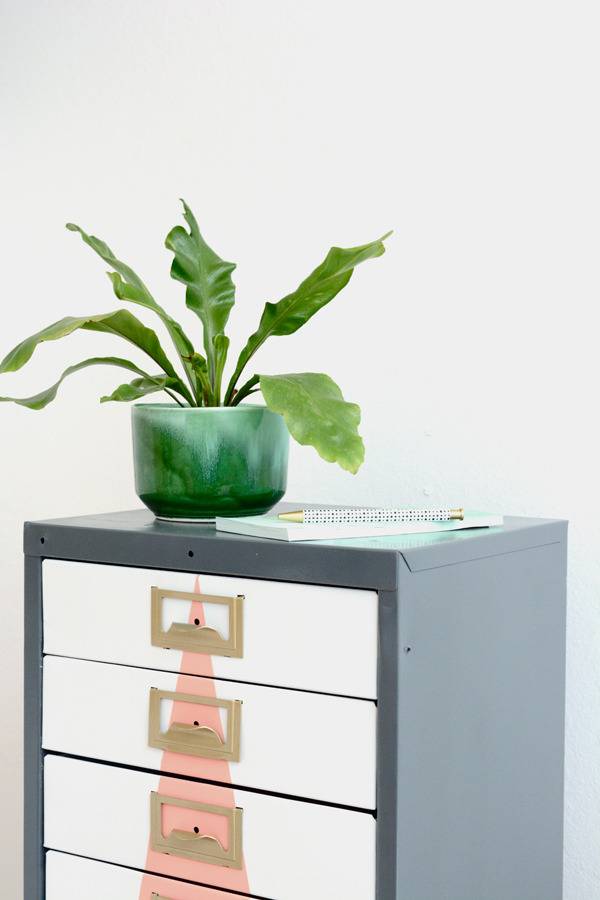 A multi drawered grey and white dresser with a pink triangle on the front and a green plant in a green planter on top of it.