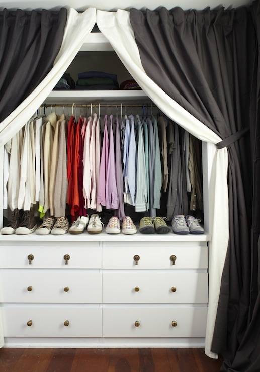 A black and white curtained tied back revealing a neat closet with clothes hung by color over shoes lined atop a white dresser.