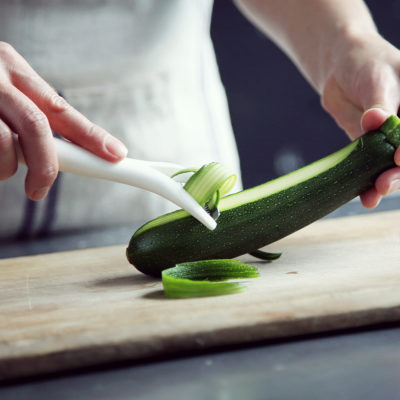 Hands using a paring tool to remove outer skin from a green zucchini on a cutting board.