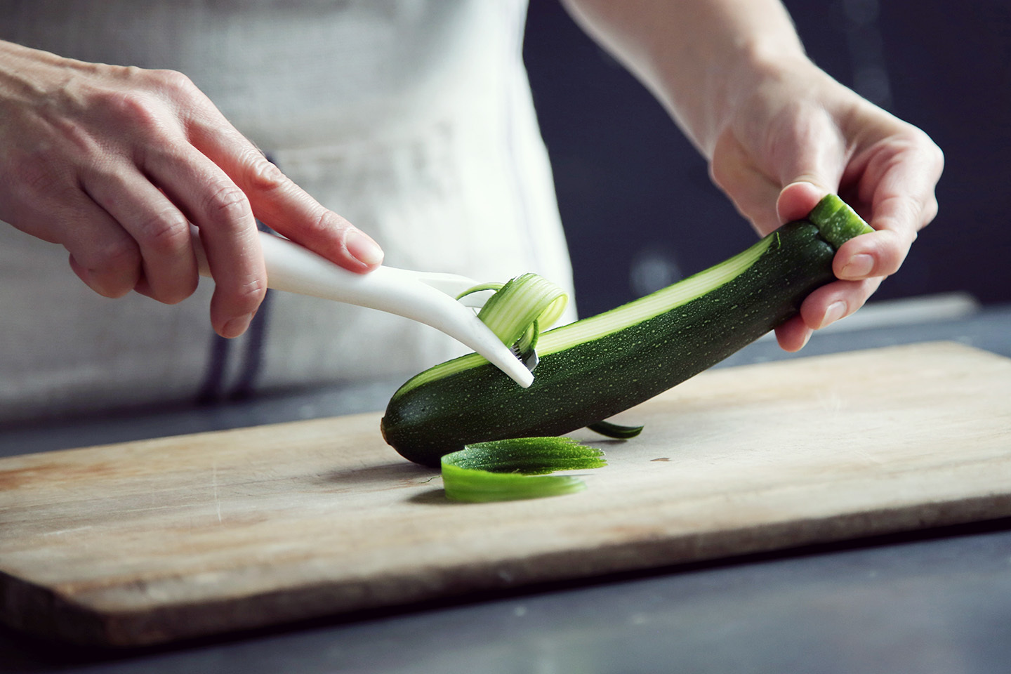 Hands using a paring tool to remove outer skin from a green zucchini on a cutting board.