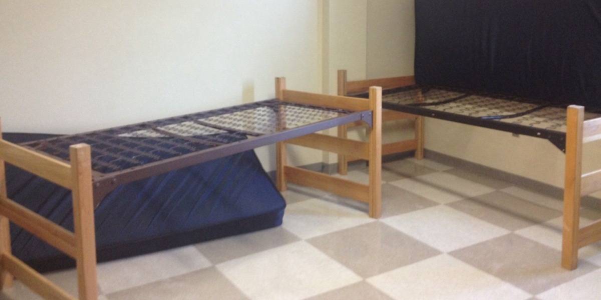 Dorm room with two simple single beds without mattresses on top and checkerboard floor.