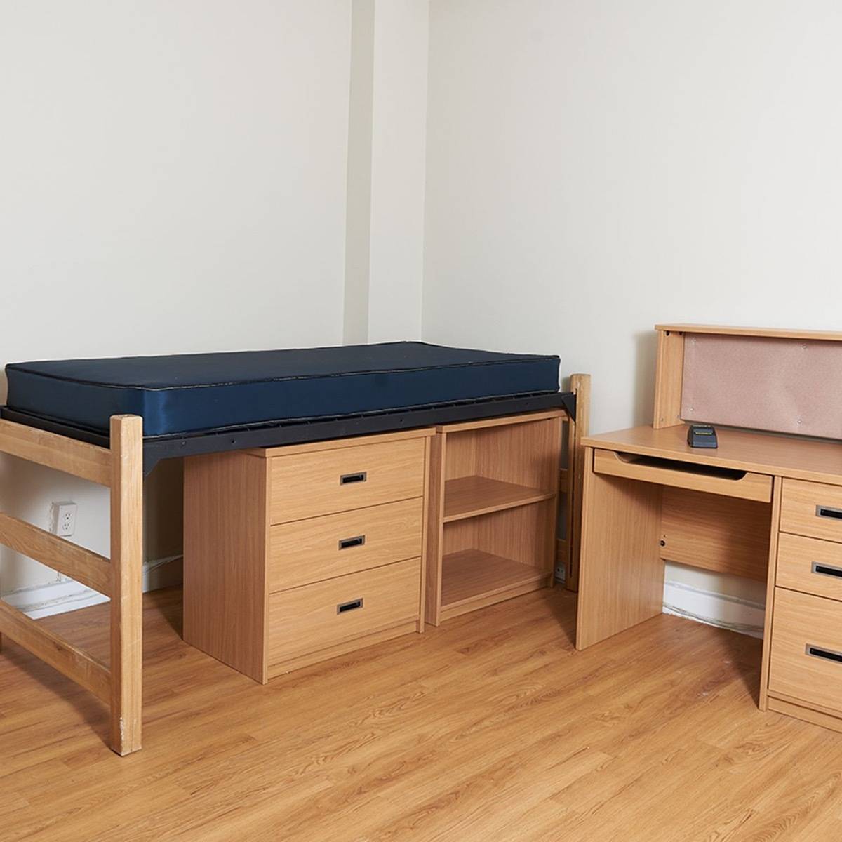 Undecorated dorm room containing a single bed with under-bed storage and a matching desk.