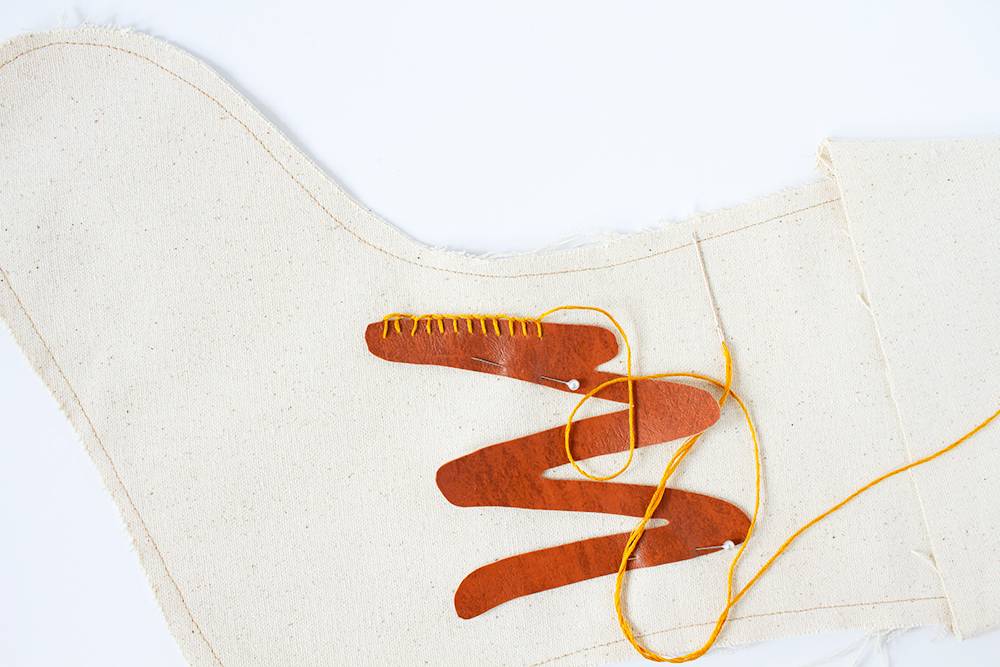 Leather monogram stocking being stitched together with yellow string on top of a white table.