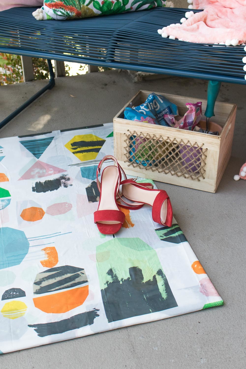 A pair of red heels laying on a multicolored blanket next to a box of pink and blue material on the pavement below a blue bench.